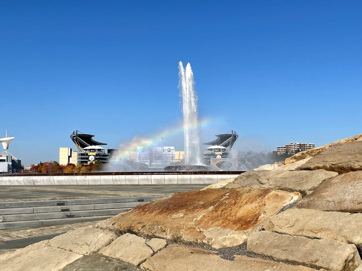 A rainbow arching over the Point State Park fountain in Pittsburgh with Acrisure Stadium visible in the background under a clear blue sky.
