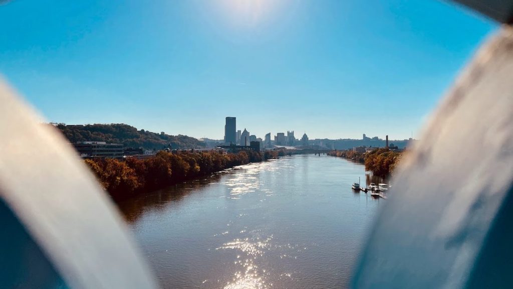 Panoramic view of the Pittsburgh skyline and the Allegheny River framed through the circular architecture of the Herr’s Island footbridge on a sunny day with fall foliage.