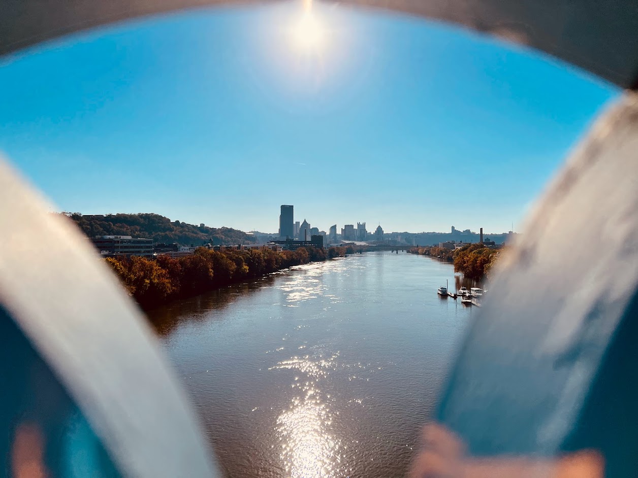 Panoramic view of the Pittsburgh skyline and the Allegheny River framed through the circular architecture of the Herr’s Island footbridge on a sunny day with fall foliage.