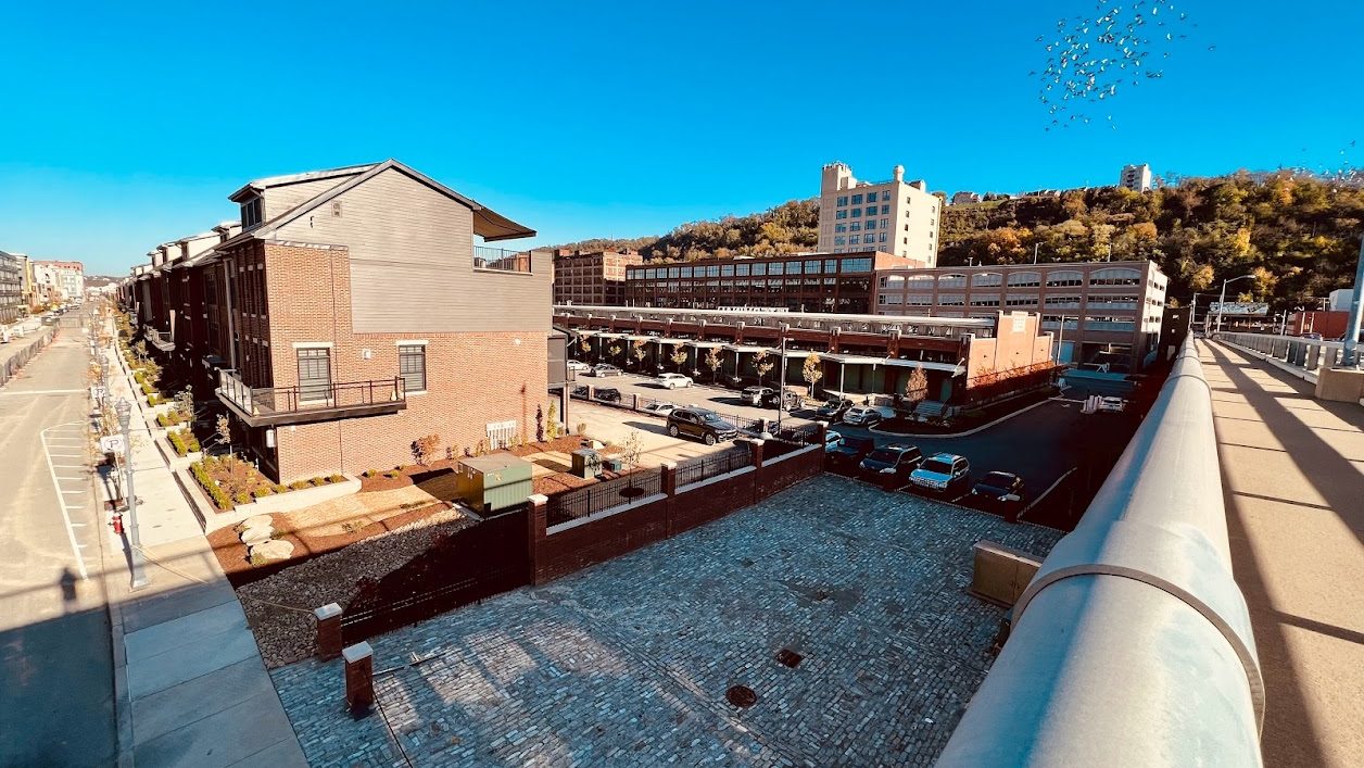 Wide-angle view of the Pittsburgh Strip District featuring historic cobblestone streets, modern brick townhomes, and the lush hills of Polish Hill in the background.