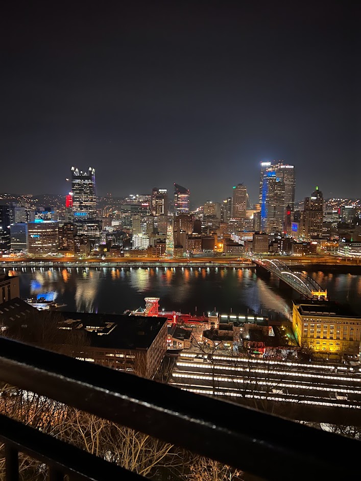 Nighttime panoramic view of the Pittsburgh skyline featuring illuminated skyscrapers, the Golden Triangle, and city lights reflecting on the Monongahela River as seen from Emerald View Park.